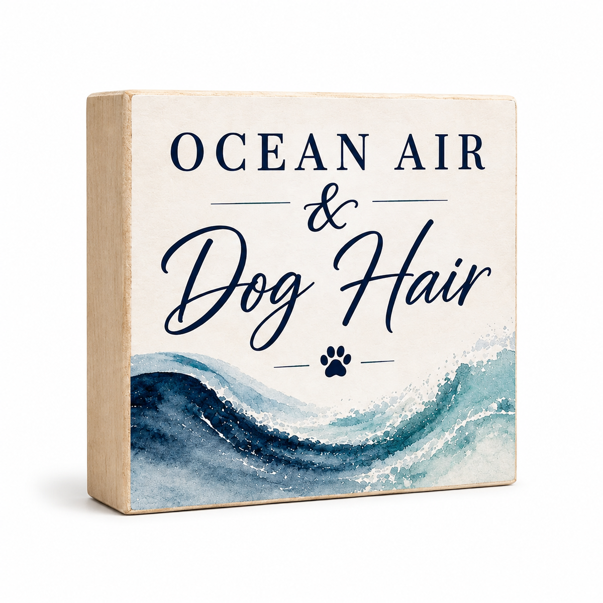 A square decorative wooden block sign with a light wood side and a cream front panel. The front reads 'OCEAN AIR & Dog Hair' in navy text above a blue and teal watercolor wave design with a small navy paw print.