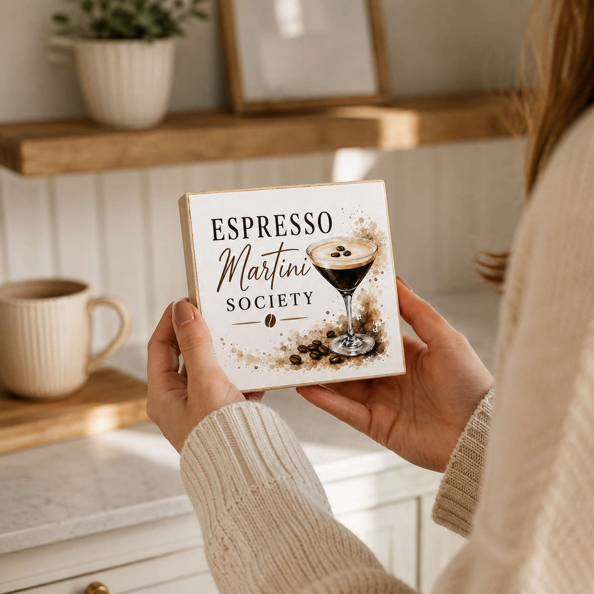 A person in a cream knit sweater holds a square decorative coaster featuring the text 'ESPRESSO Martini SOCIETY' and an illustration of an espresso martini glass with coffee beans, set against a warm, softly blurred kitchen background.