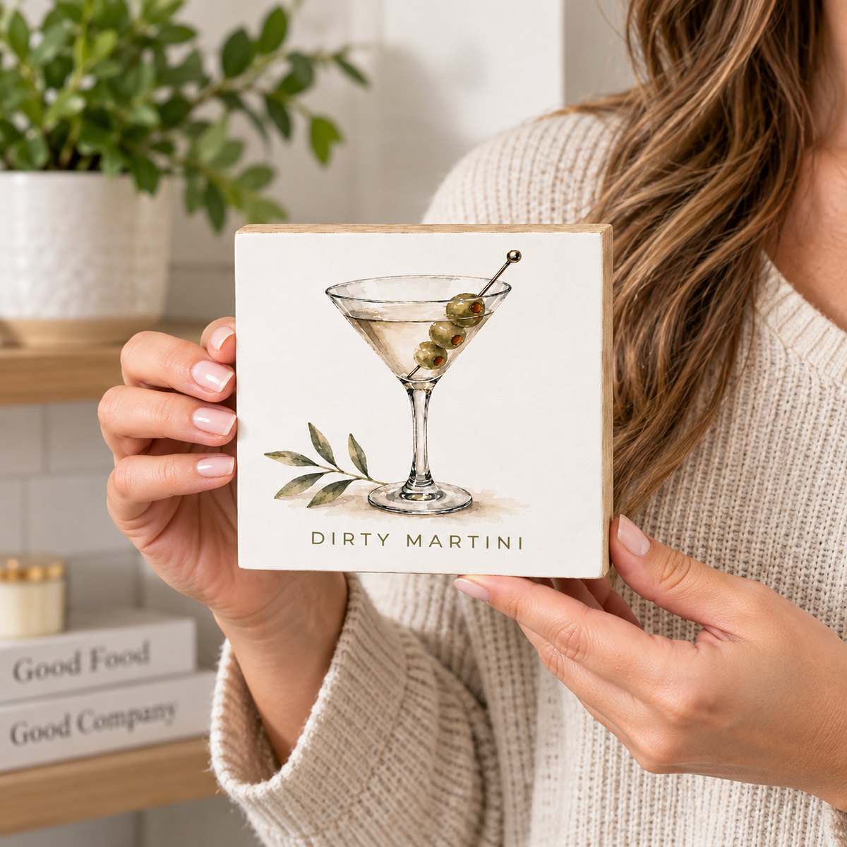 A person in a beige knit sweater holds a square ceramic coaster featuring an illustration of a martini glass with olives and the text 'DIRTY MARTINI'. The background shows a softly blurred kitchen setting with a plant and books.