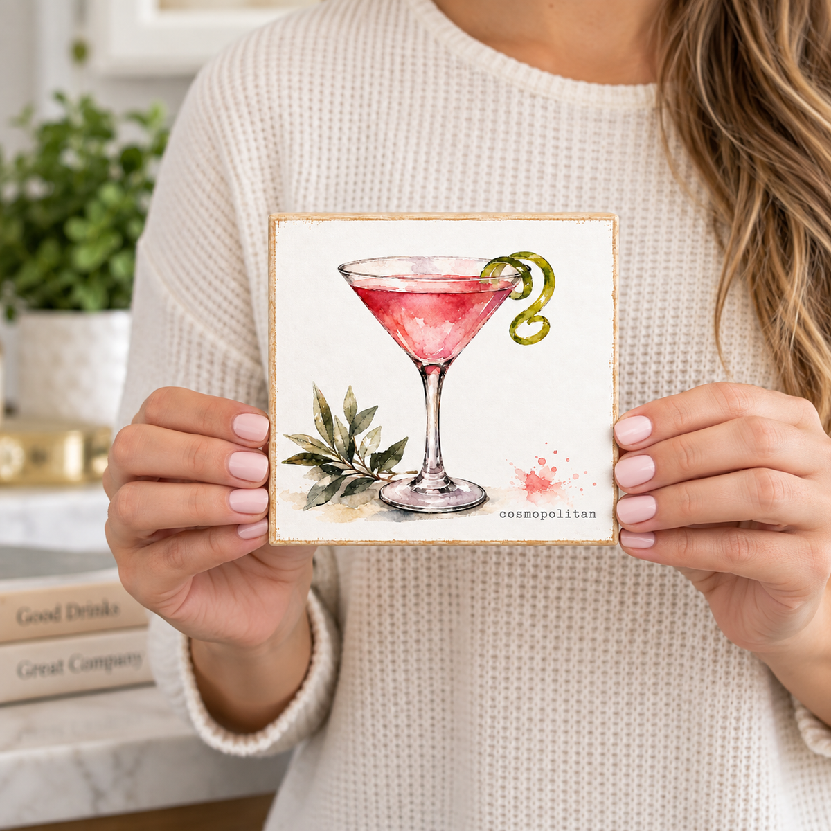 A woman in a cream knit sweater holds a square coaster featuring a watercolor illustration of a pink martini cocktail with a green twist and olive branches. The word 'cosmopolitan' is printed at the bottom.