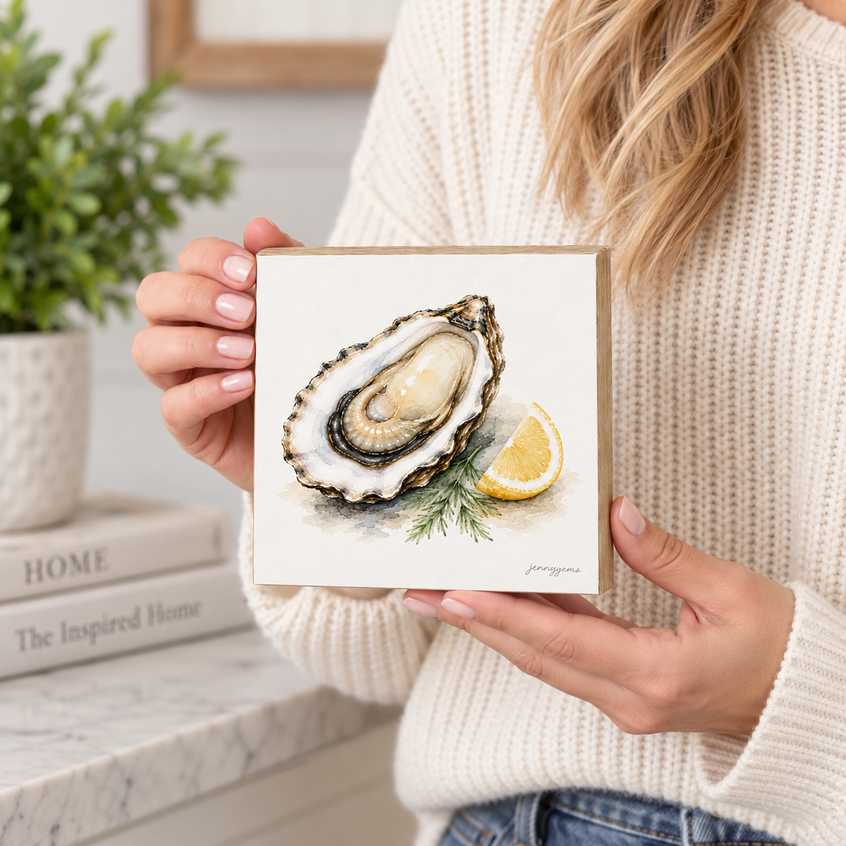 A person in a cream knit sweater holds a square art print featuring a painted oyster shell with a lemon wedge and green herb sprig. The scene is styled indoors with a plant and books in the background.