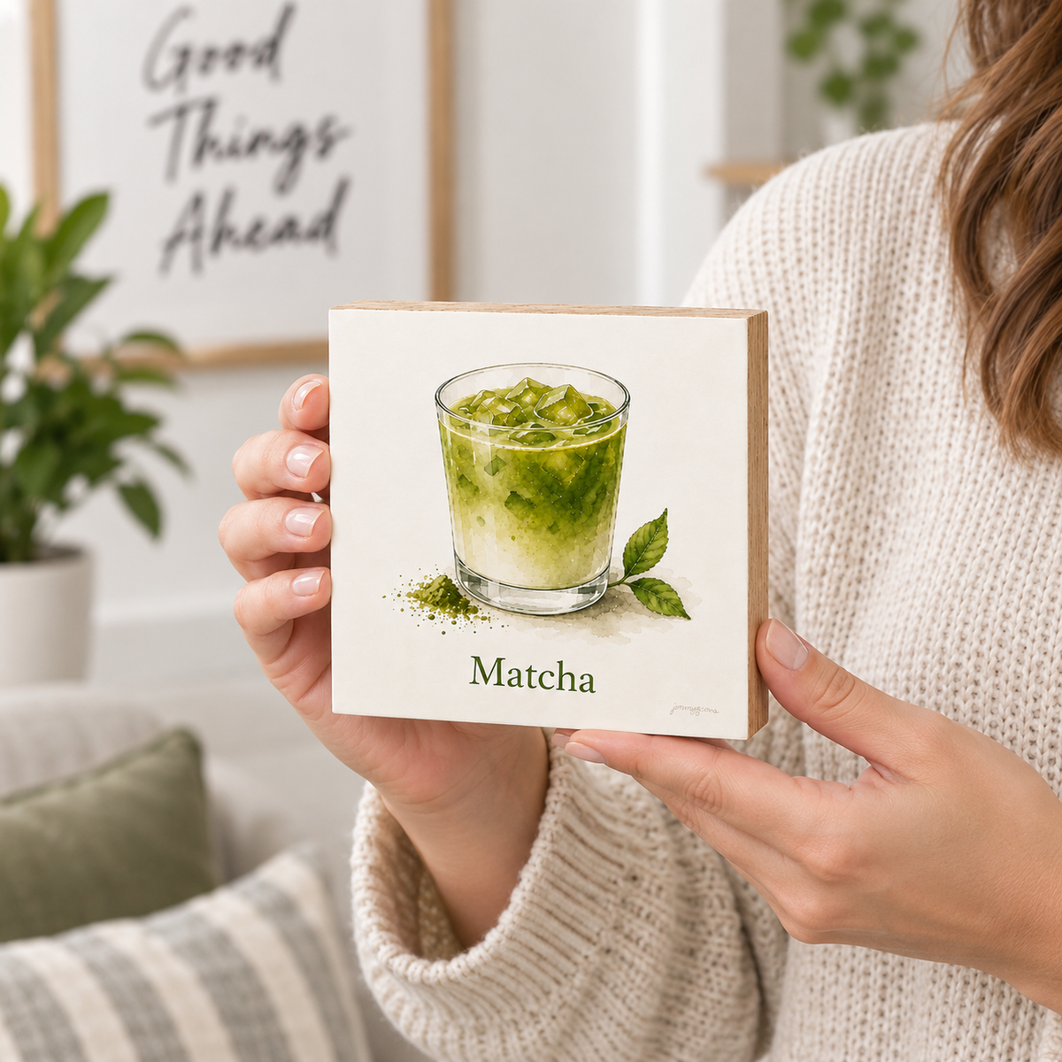 A person in a cream knit sweater holds a square white card or coaster featuring a painted matcha drink in a glass with the word "Matcha" below. The background is a softly blurred living room with a framed quote and plants.
