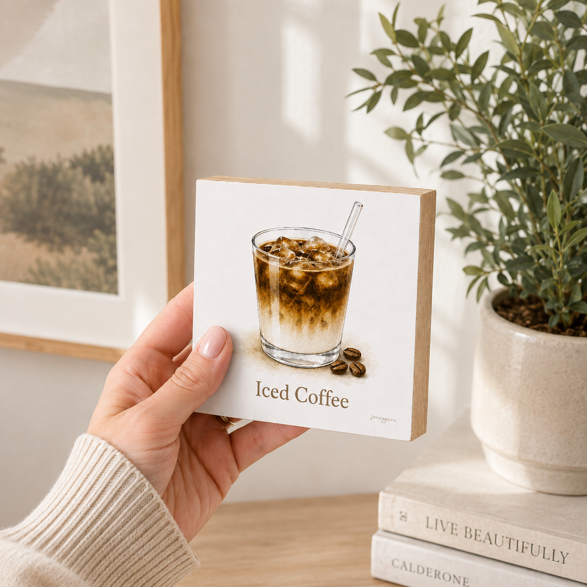 A hand holds a square white coaster featuring an illustration of an iced coffee glass with the text 'Iced Coffee' on a light wood background, with a potted plant and books in the softly lit room behind it.