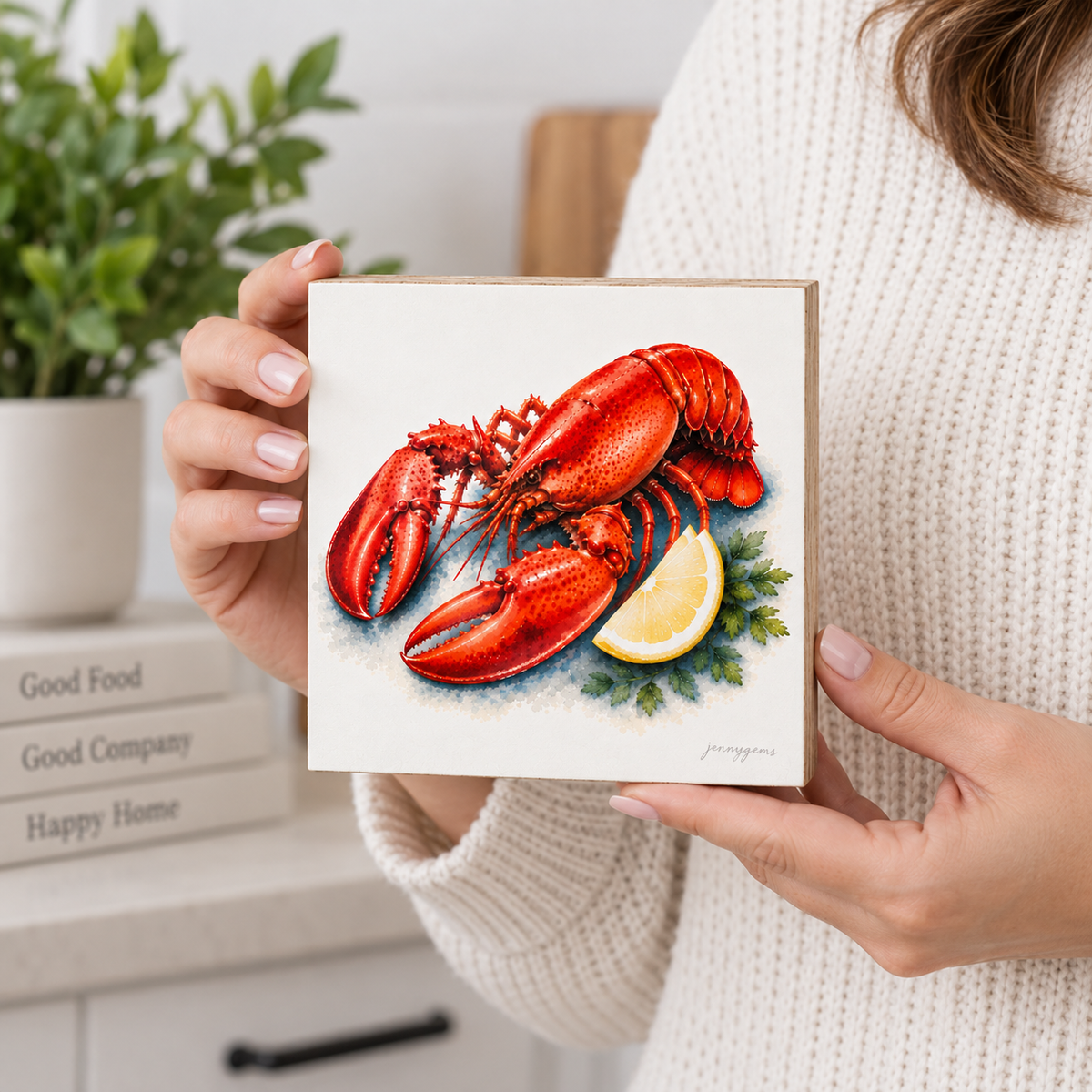 A person in a cream knit sweater holds a square canvas print featuring a detailed illustration of a red lobster with a lemon wedge and green herbs on a white background. The setting is a bright, modern kitchen with a blurred plant and books in the background.