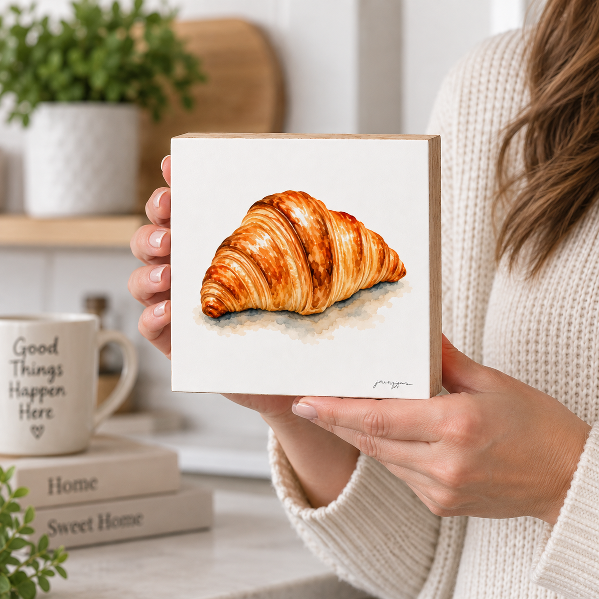 A person in a cream knit sweater holds a square white art print or tile featuring a painted golden-brown croissant. The background shows a cozy kitchen or cafe setting with a mug on stacked books and a plant.