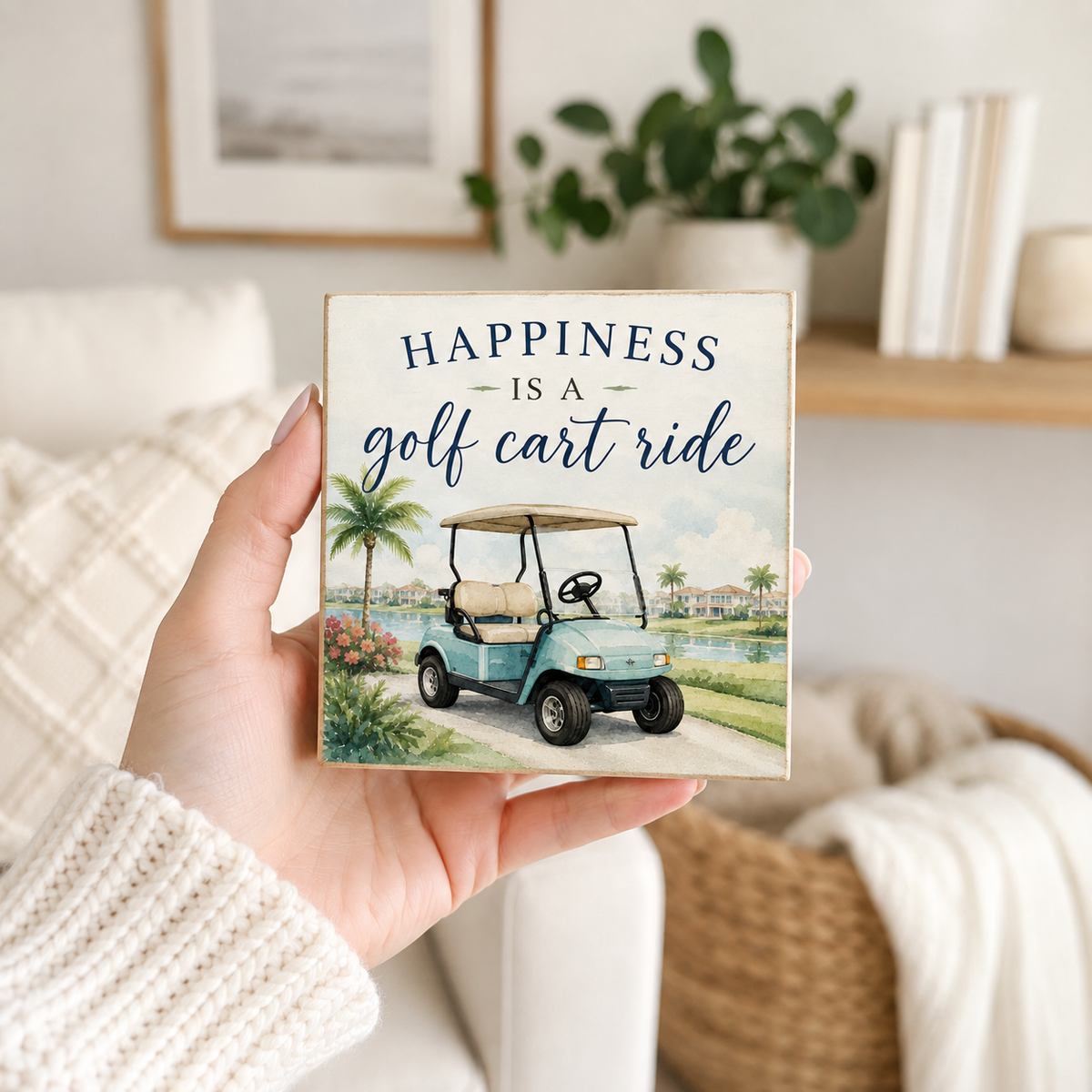 A hand holds a square decorative sign featuring the phrase 'HAPPINESS IS A golf cart ride' above an illustration of a light blue golf cart on a golf course. The background shows a cozy, softly blurred living room setting with a sofa, books, and a plant.
