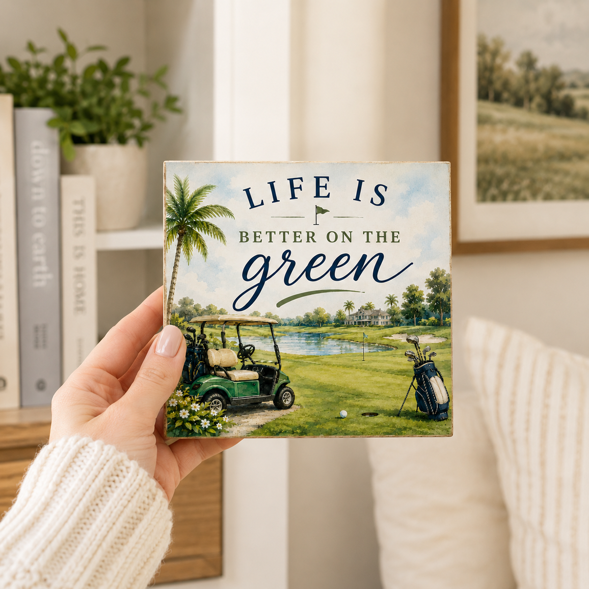 A hand wearing a cream knit sweater holds a square decorative plaque featuring a golf course scene with the text 'LIFE IS BETTER ON THE green.' The background shows a softly blurred living room interior with a sofa, framed wall art, books, and a plant.