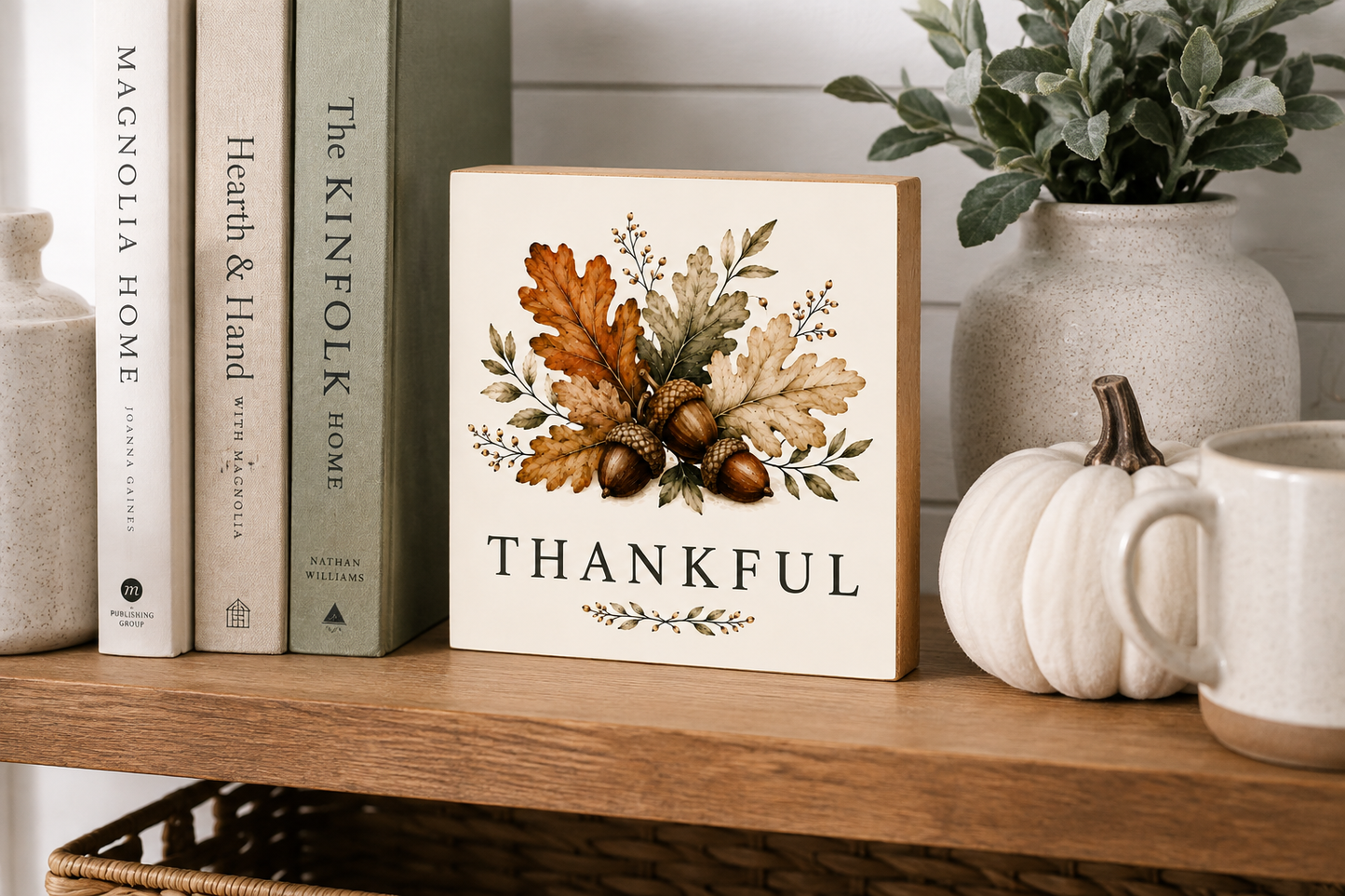 A square decorative sign with a cream background and a botanical autumn design featuring oak leaves, acorns, and the word 'THANKFUL' in black serif text. The sign is styled on a wooden shelf next to books, a ceramic pumpkin, a mug, and a potted plant.