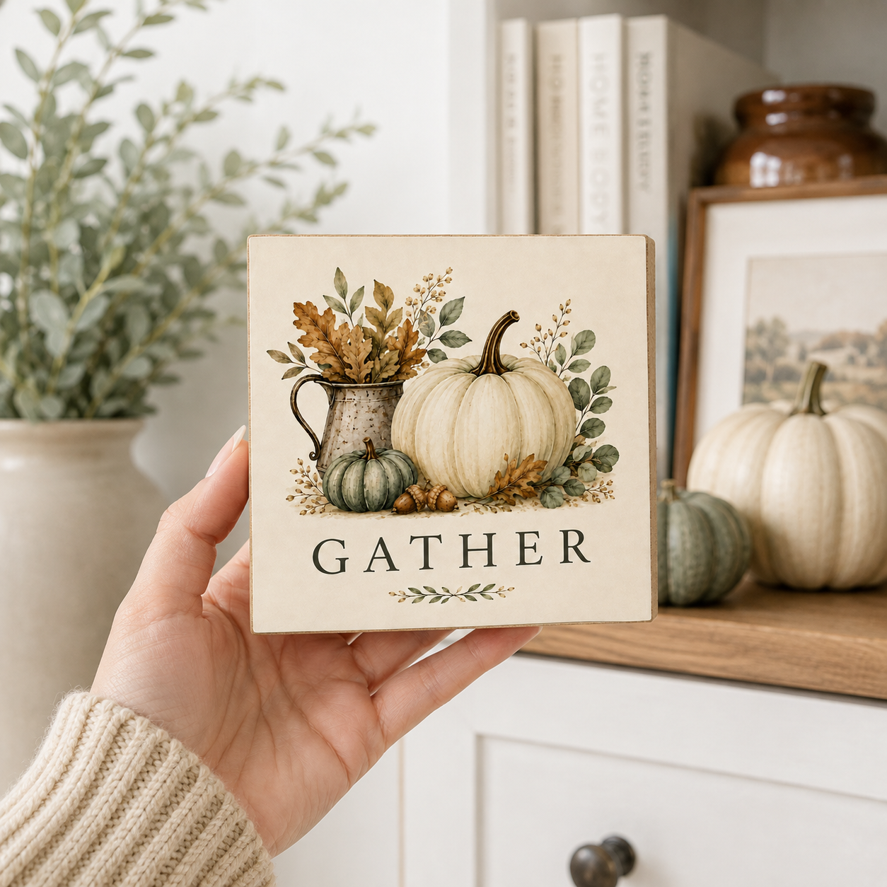 A hand holds a square decorative tile featuring a fall harvest illustration with pumpkins, leaves, and the word “GATHER” in black serif text. The background shows a cozy home interior with books, framed art, and more pumpkins on a shelf.