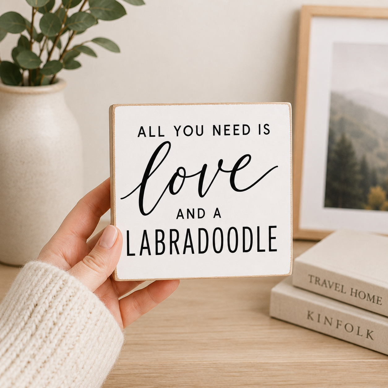 A hand wearing a cream knit sweater holds a square white wooden sign with black text that reads, 'ALL YOU NEED IS love AND A LABRADOODLE.' The scene is a styled indoor tabletop setup with a vase of greenery, a framed picture, and stacked books in the background.