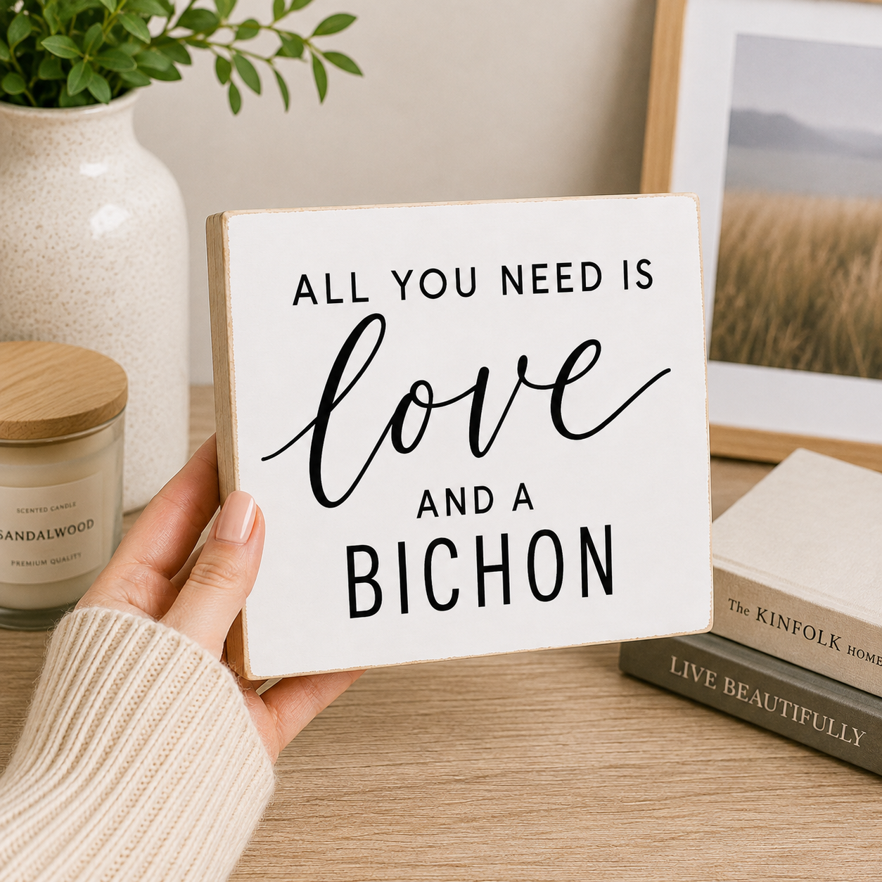 A hand wearing a cream knit sweater holds a square white wooden sign with black text that reads 'ALL YOU NEED IS love AND A BICHON'. The background shows a styled home interior with a candle, a plant, a framed picture, and books on a wooden table.