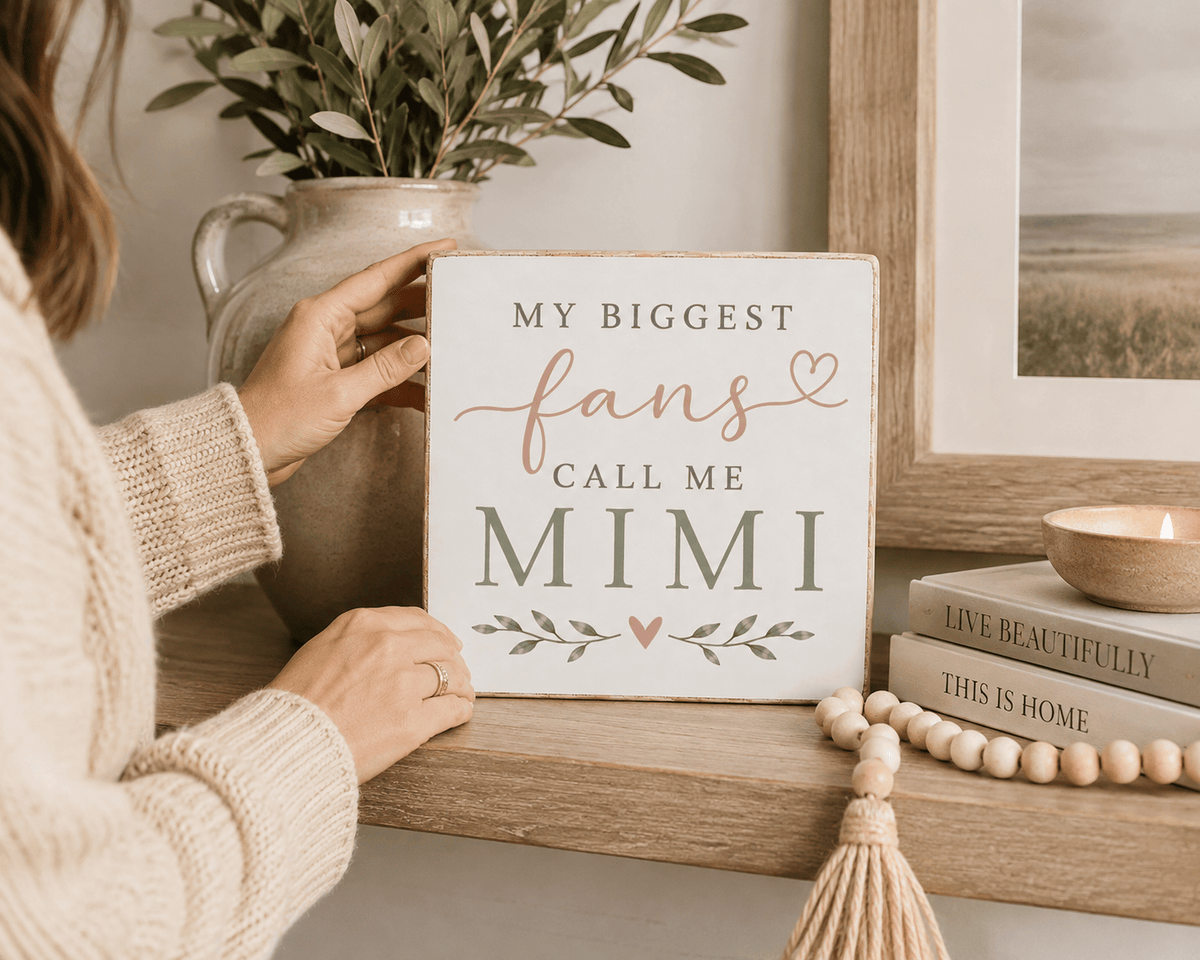 A person in a cream knit sweater holds a square white decorative sign that reads 'MY BIGGEST fans CALL ME MIMI' with a blush pink script 'fans' and small floral accents. The sign is styled on a wooden shelf beside a ceramic vase with greenery, a framed print, books, and a candle.