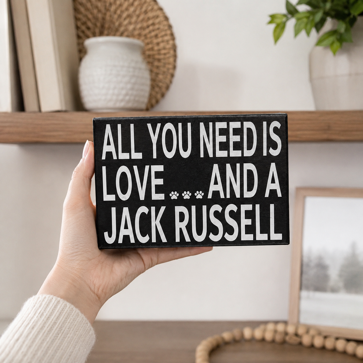 A hand holds a black rectangular wooden sign with white text that reads 'ALL YOU NEED IS LOVE.