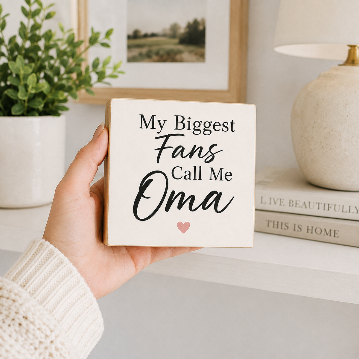 A hand wearing a cream knit sweater holds a square wooden sign that reads 'My Biggest Fans Call Me Oma' with a small pink heart. The background shows a styled home interior with a potted plant, framed picture, lamp, and books.