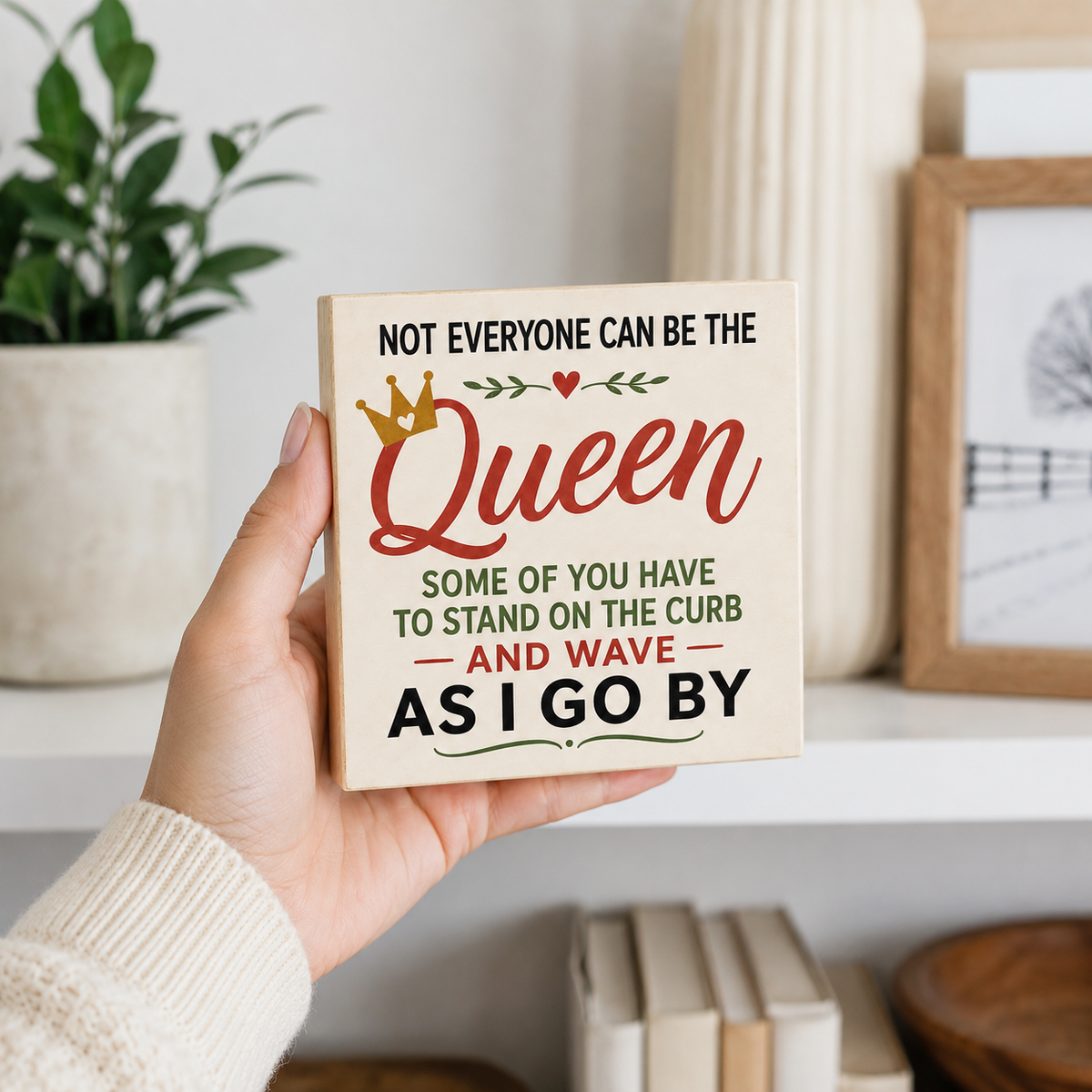 A hand holding a square decorative sign that reads 'NOT EVERYONE CAN BE THE Queen SOME OF YOU HAVE TO STAND ON THE CURB AND WAVE AS I GO BY' in black and red lettering with green accents, set against a softly blurred home decor background.