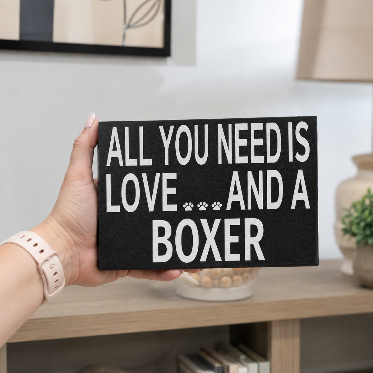 A hand holds a black rectangular sign with bold white text reading 'ALL YOU NEED IS LOVE *** AND A BOXER' against a softly blurred indoor background.