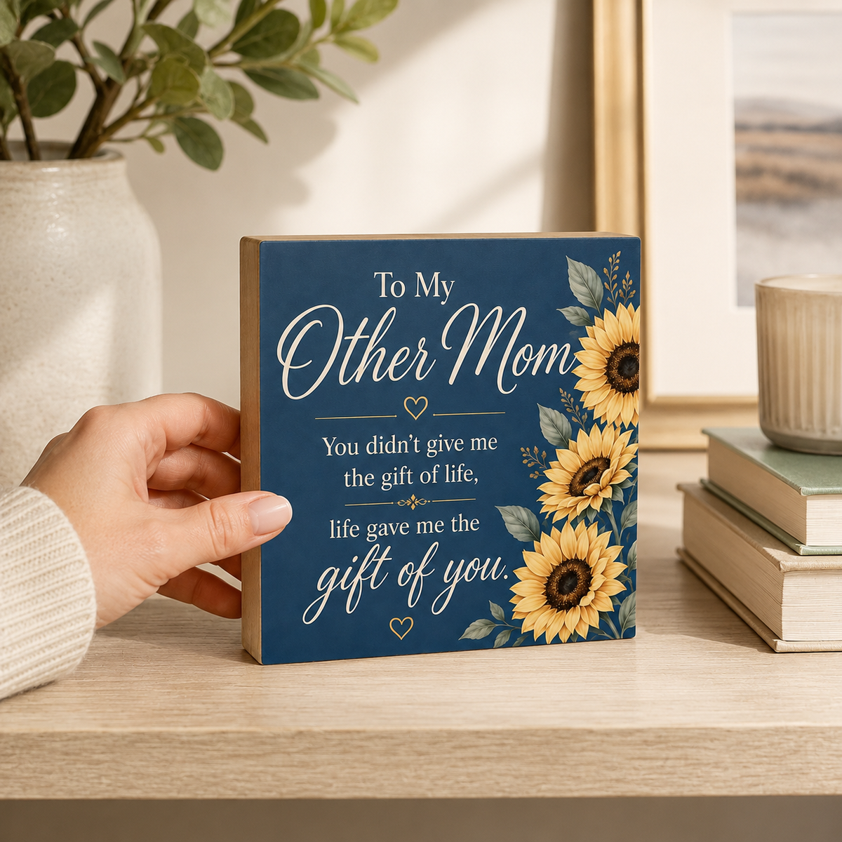 A hand holds a square wooden sign with a navy blue background, white cursive text reading 'To My Other Mom,' and yellow sunflower illustrations on the right side, set on a light wood shelf in a home interior.