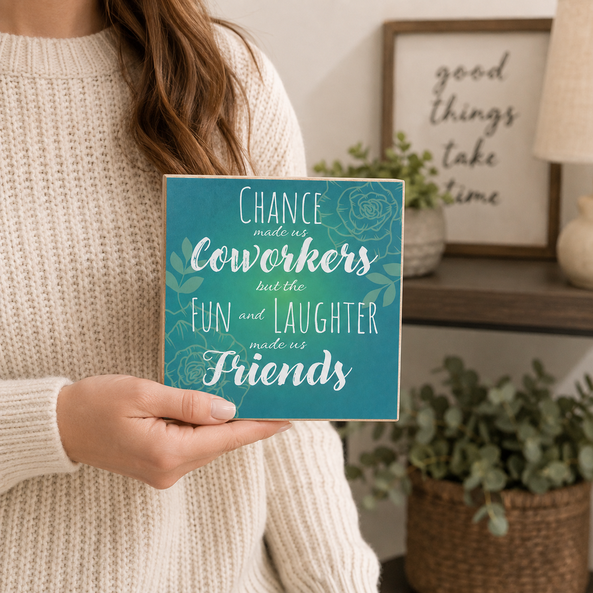 A person in a cream knit sweater holds a square teal sign with white text that reads, 'Chance made us coworkers but the fun and laughter made us friends.' The background shows a softly styled indoor room with framed wall art, a lamp, and potted plants.