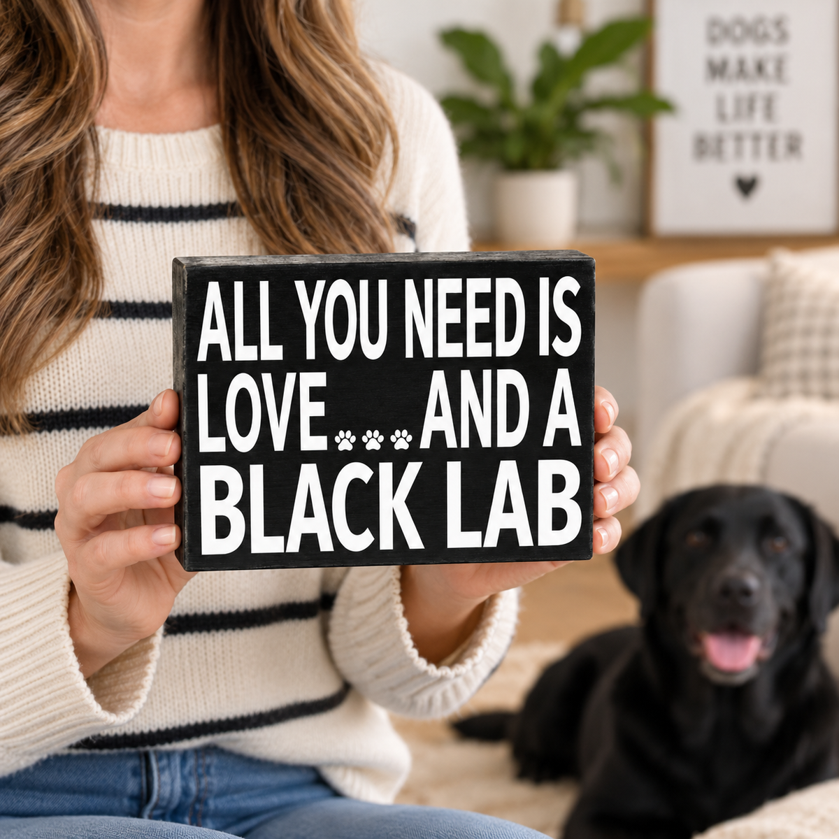 A woman in a cream sweater holds a black wooden sign that reads 'ALL YOU NEED IS LOVE.