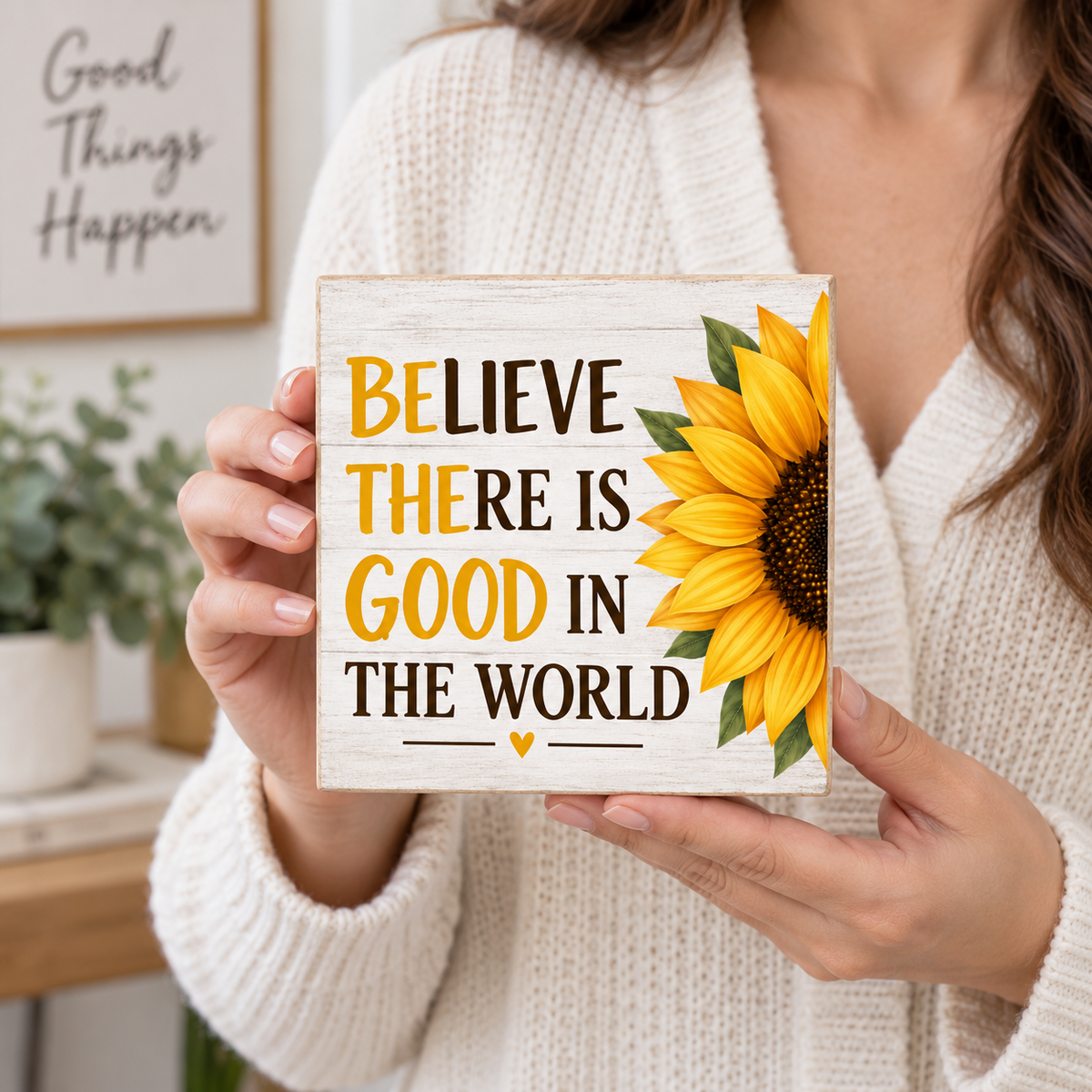 A woman in a cream knit sweater holds a square wooden sign with the phrase “BELIEVE THERE IS GOOD IN THE WORLD” in yellow and dark brown lettering, next to a large yellow sunflower. The background shows a softly blurred home interior with a framed sign that reads “Good Things Happen.