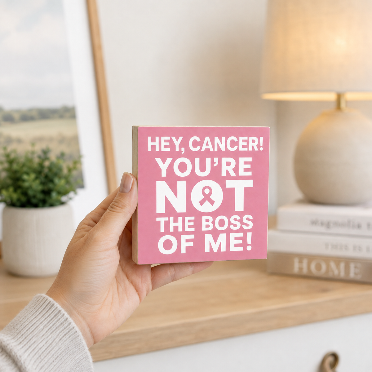 A hand holds a square pink sign with the text 'HEY, CANCER! YOU'RE NOT THE BOSS OF ME!' in white lettering with a pink ribbon. The background shows a softly blurred home interior with a lamp, books, and a plant.