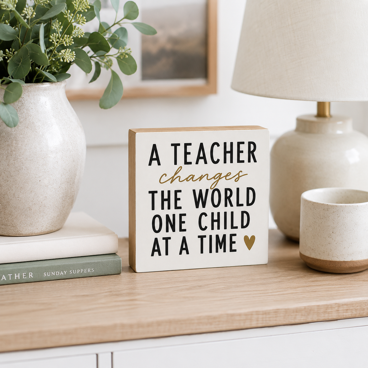 A square wooden sign with the quote 'A TEACHER changes THE WORLD ONE CHILD AT A TIME' in black and gold lettering, styled on a light wood table next to a vase with greenery, a lamp, and a ceramic cup.