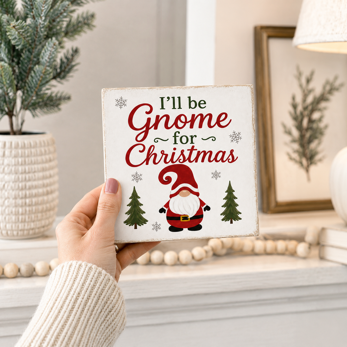 A hand holds a square white decorative sign that reads “I’ll be Gnome for Christmas” with a gnome illustration and small pine trees, set against a cozy indoor holiday background.