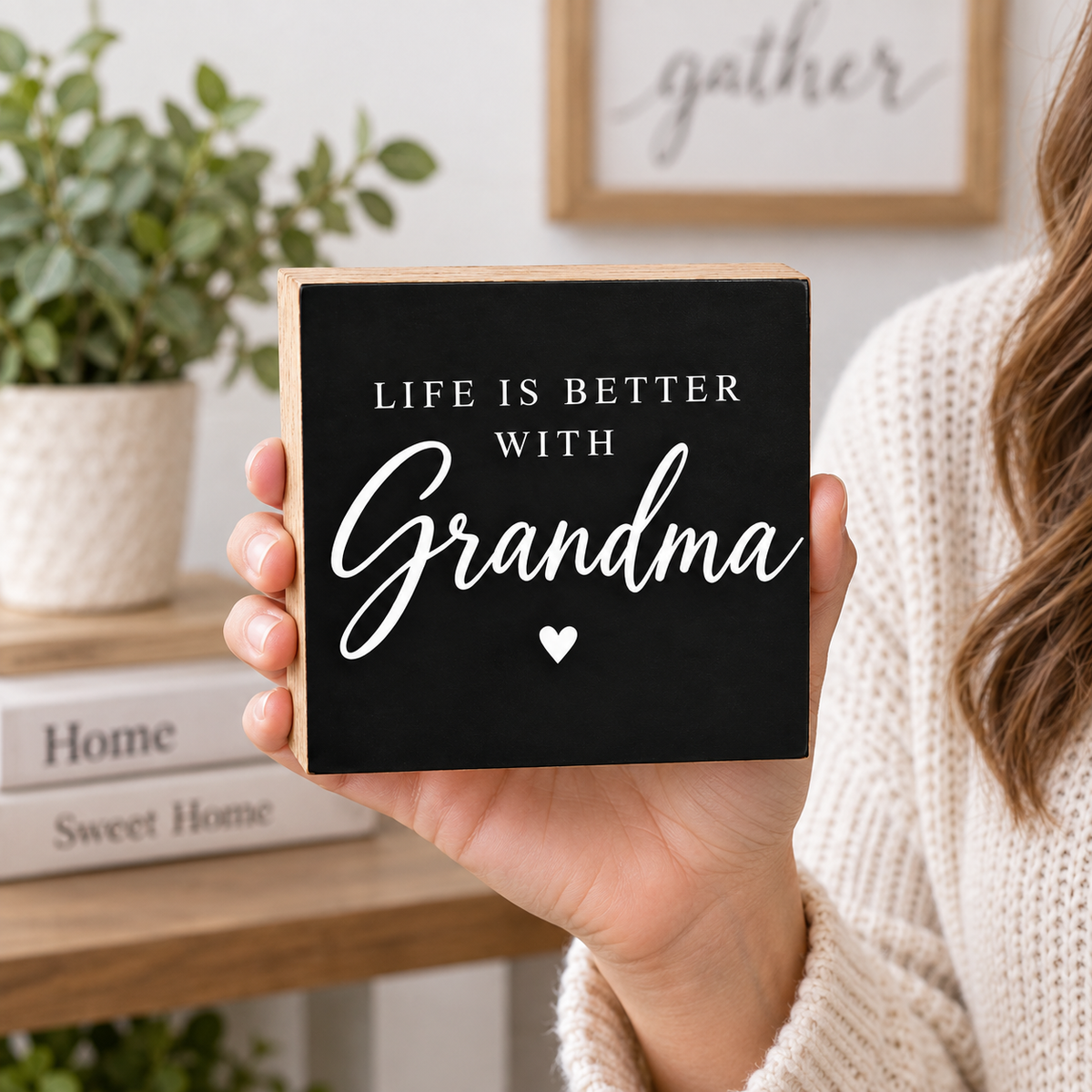 A person in a cream knit sweater holds a square black wooden sign with natural wood edges. The sign reads “LIFE IS BETTER WITH Grandma” in white lettering with a small heart below.