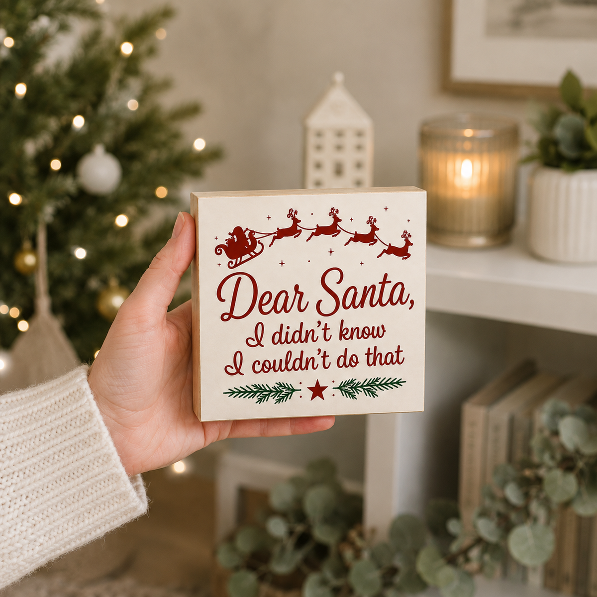 A hand in a white knit sweater holds a square wooden sign that reads 'Dear Santa, I didn't know I couldn't do that' with red Santa and reindeer graphics and green pine accents. The background shows a cozy holiday scene with a lit Christmas tree, candles, and plants.