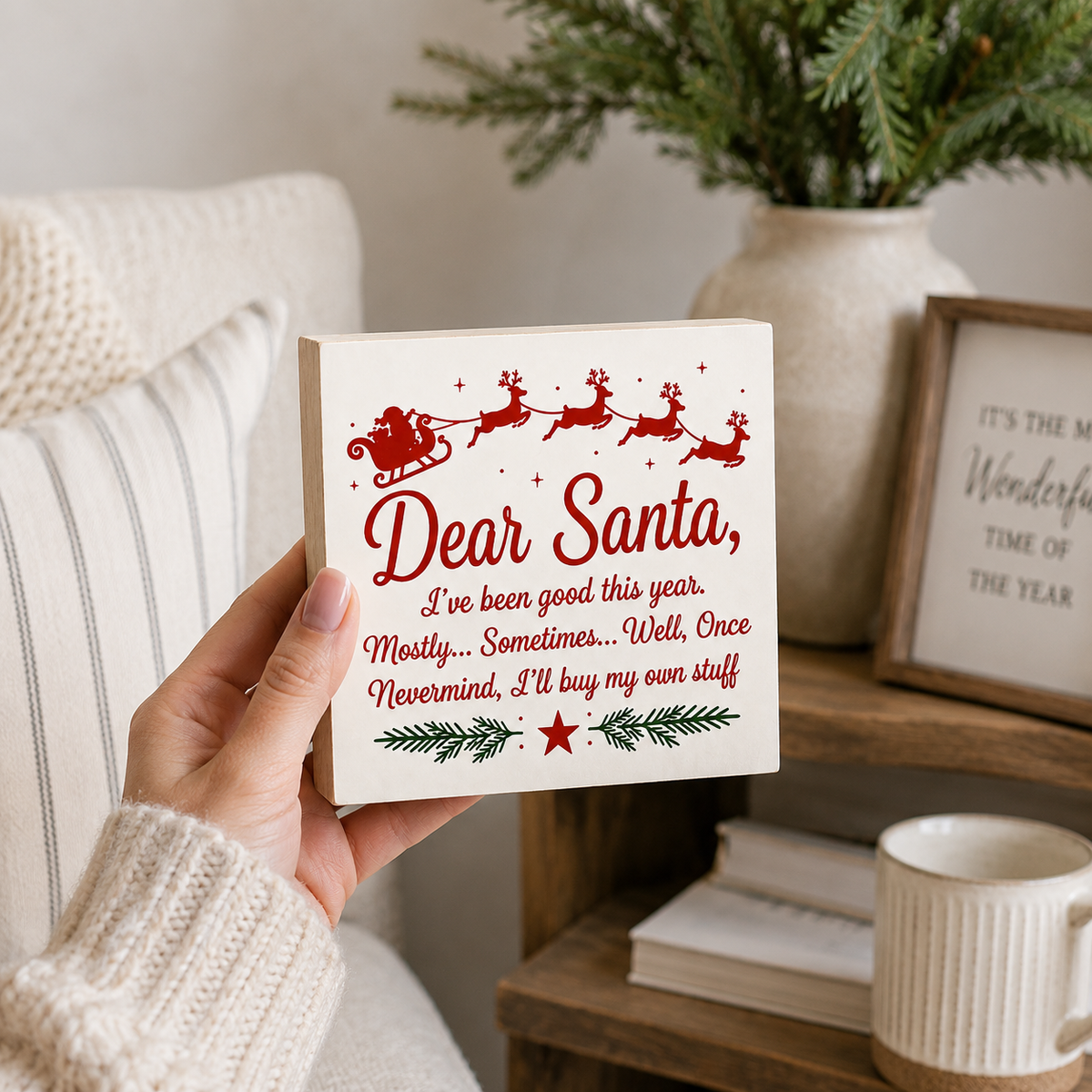 A hand in a cream knit sweater holds a square white decorative sign with a red and green Christmas message to Santa. The sign features a red sleigh and reindeer illustration at the top and a red star near the bottom.
