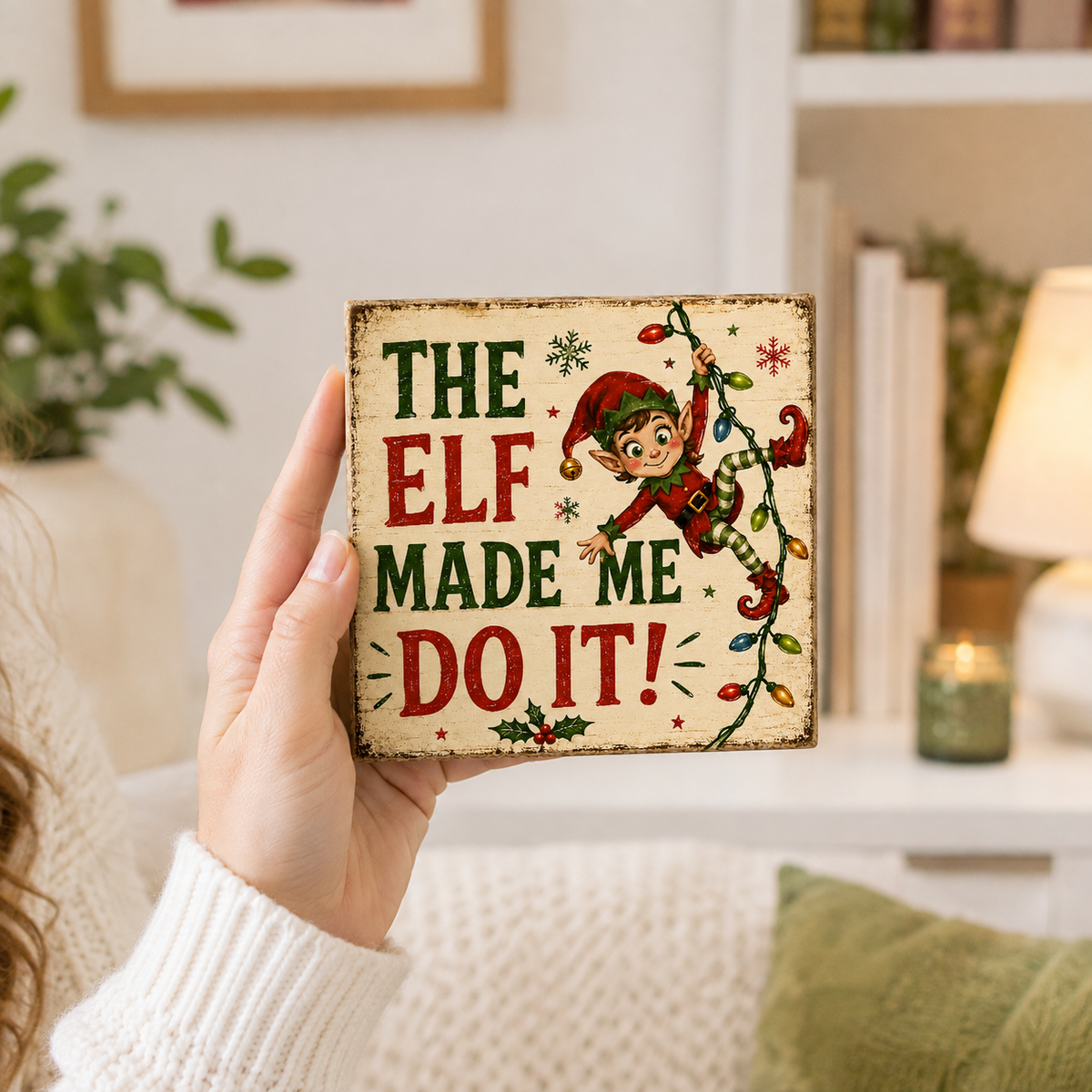 A hand holds a square decorative sign with a distressed cream background, featuring the text 'THE ELF MADE ME DO IT!' in red and green lettering, alongside an illustration of a Christmas elf climbing a string of multicolored lights. The background shows a cozy, softly blurred living room setting with a lamp, books, and a plant.