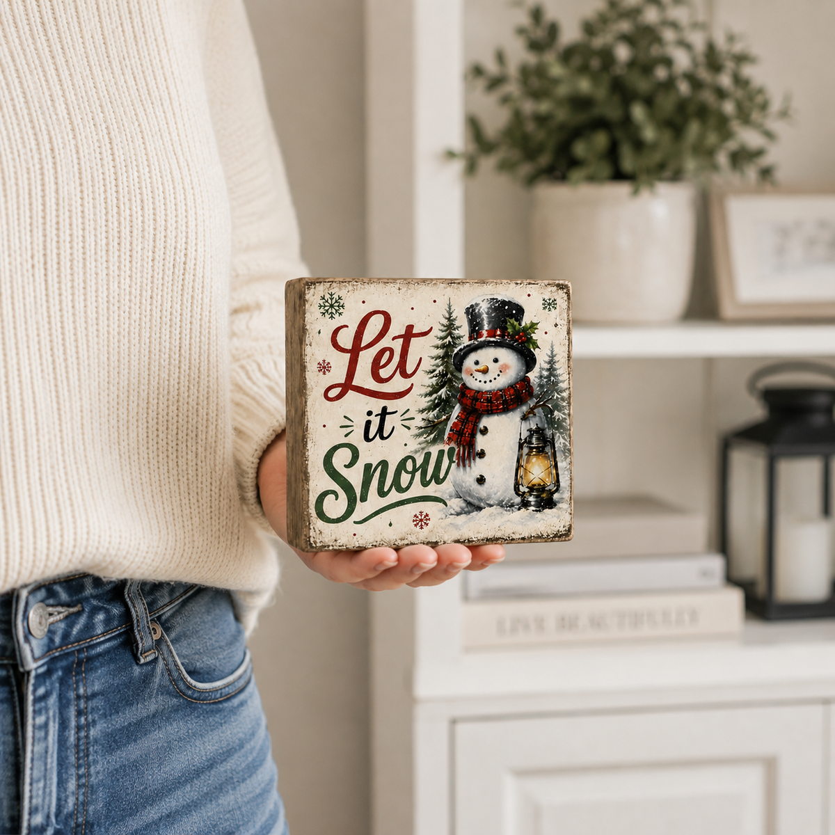 A person in a cream knit sweater and blue jeans holds a square decorative sign that reads “Let it Snow” with a winter snowman illustration. The background shows a softly blurred indoor room with shelves, a plant, and a lantern.