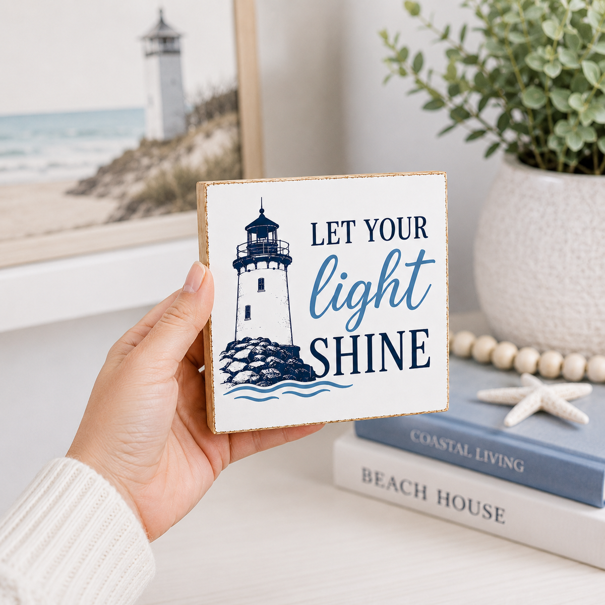 A hand holds a square white decorative sign with a lighthouse illustration and the phrase “LET YOUR light SHINE” in navy and blue lettering. The sign is photographed in a styled interior setting with a framed lighthouse print, a potted plant, books, and a starfish on a light surface.