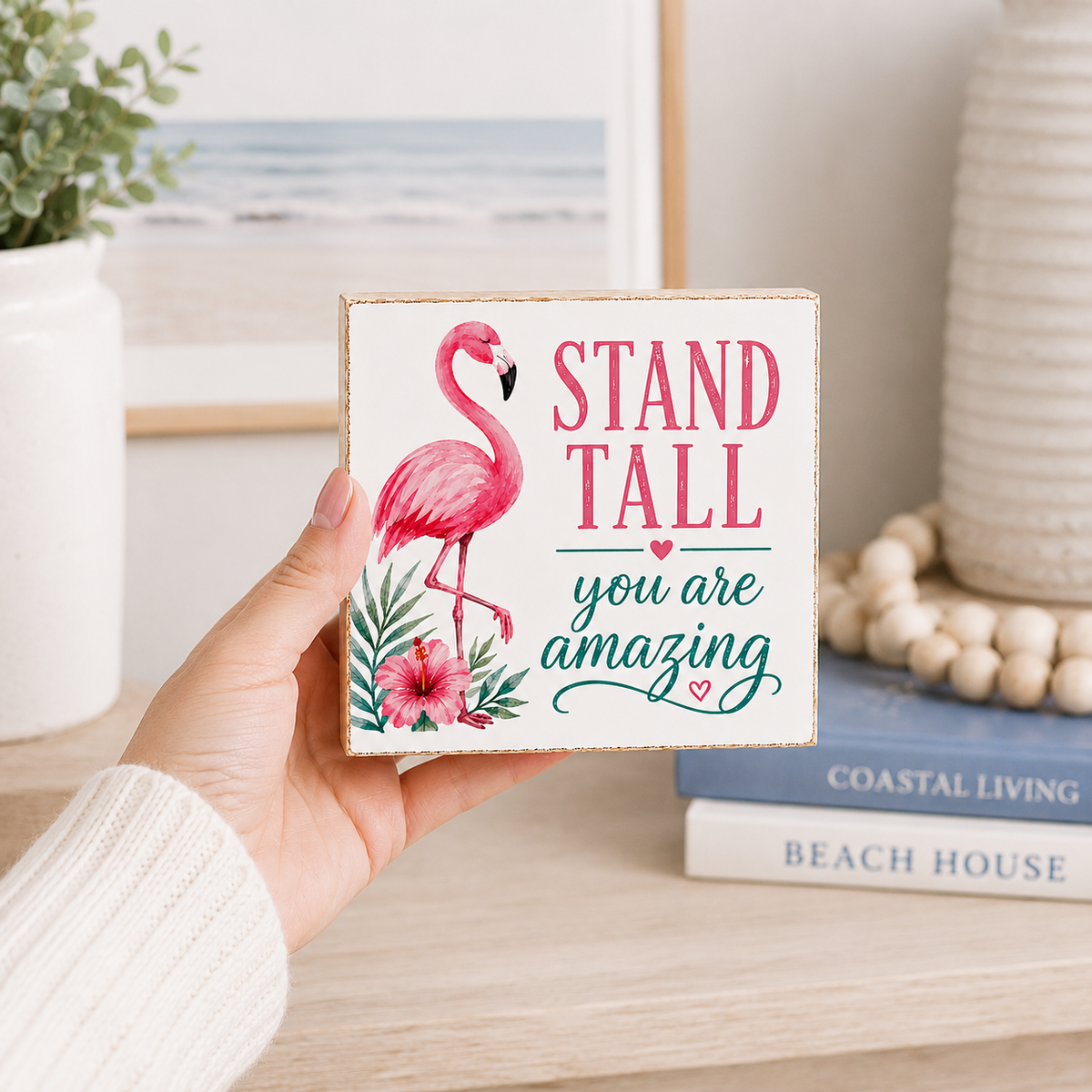 A hand holds a square decorative sign with a pink flamingo and the text 'STAND TALL you are amazing' in pink and teal lettering. The sign is styled in a beach-themed living room setting with books, a vase, and a framed print in the background.
