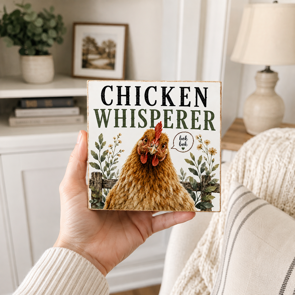 A hand holds a square decorative sign that reads 'CHICKEN WHISPERER' with a large illustrated brown hen and floral details. The scene is set in a cozy living room with a lamp, shelf, and knit blanket in the background.