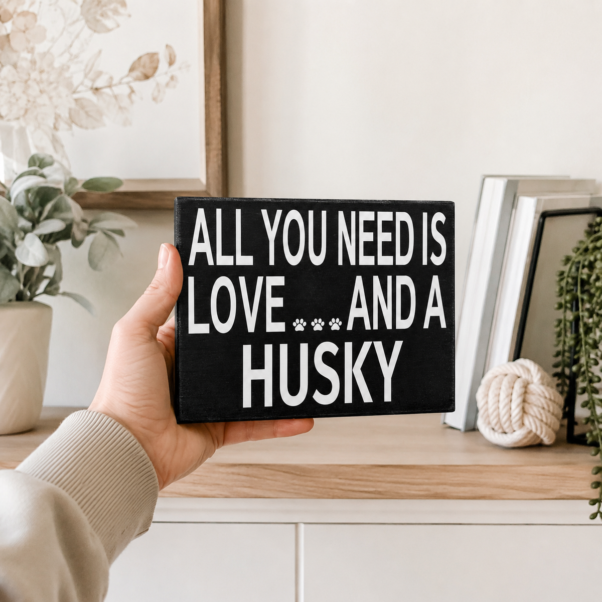 A hand holds a black rectangular sign with white text that reads 'ALL YOU NEED IS LOVE *** AND A HUSKY' with two small paw prints. The background shows a styled indoor shelf with books, a plant, and a framed picture.