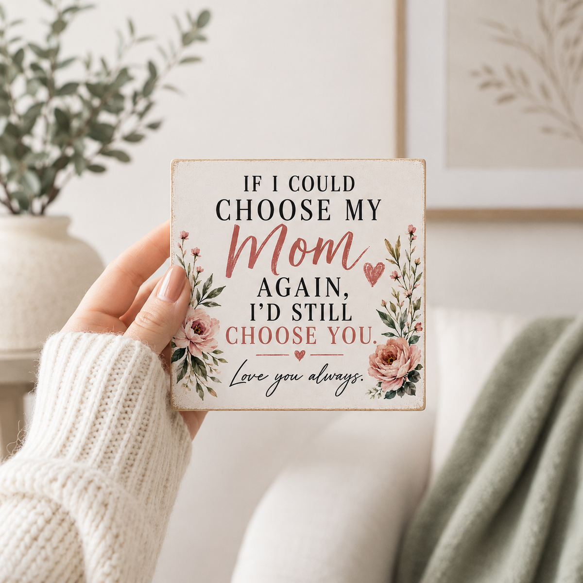 A hand in a cream knit sweater holds a square decorative sign that reads, 'IF I COULD CHOOSE MY Mom AGAIN, I'D STILL CHOOSE YOU' with floral illustrations. The background shows a softly blurred home interior with a plant, framed wall art, and a green throw blanket.