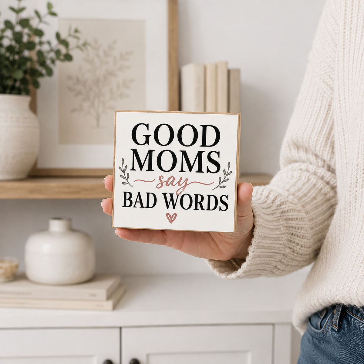 A person in a cream knit sweater holds a square wooden sign that reads 'GOOD MOMS say BAD WORDS' in black and cursive lettering, with small leaf accents and a small heart. The background shows a softly blurred home interior with shelves, books, and decor.
