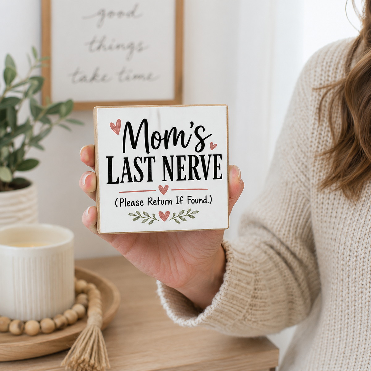 A person in a cream knit sweater holds a square wooden sign that reads 'Mom's LAST NERVE (Please Return If Found)' with small pink hearts and green leaf accents. The scene is a softly lit indoor setting with a plant, candle, and framed wall art in the background.