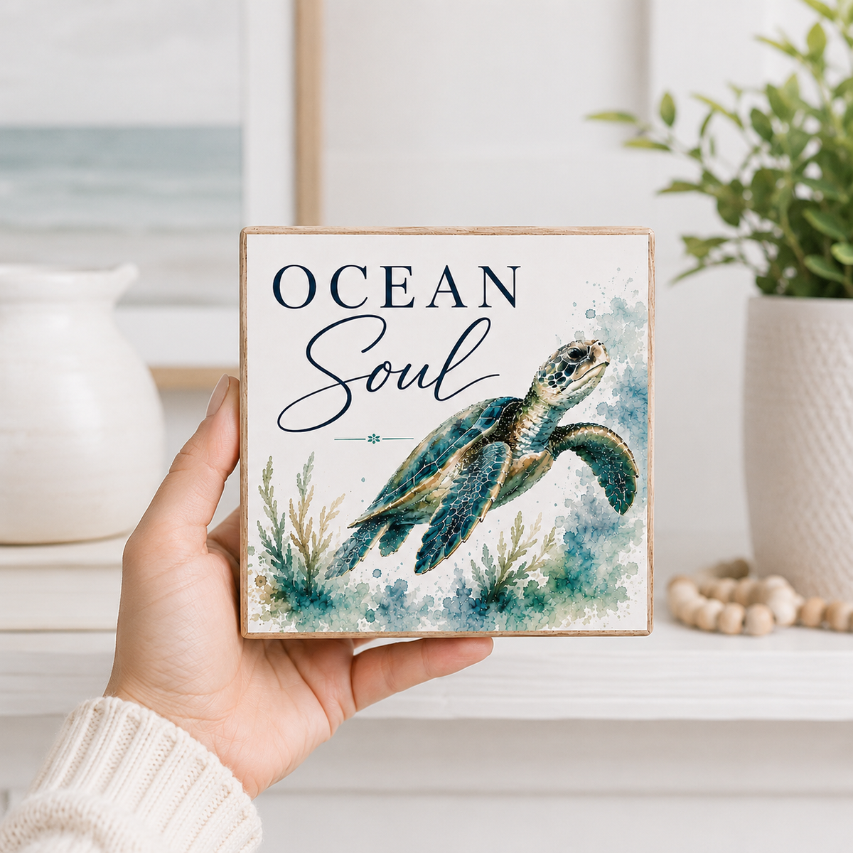 A hand holds a square wooden sign featuring a watercolor sea turtle and the words 'OCEAN Soul' in navy script, with a soft beach and plant background.