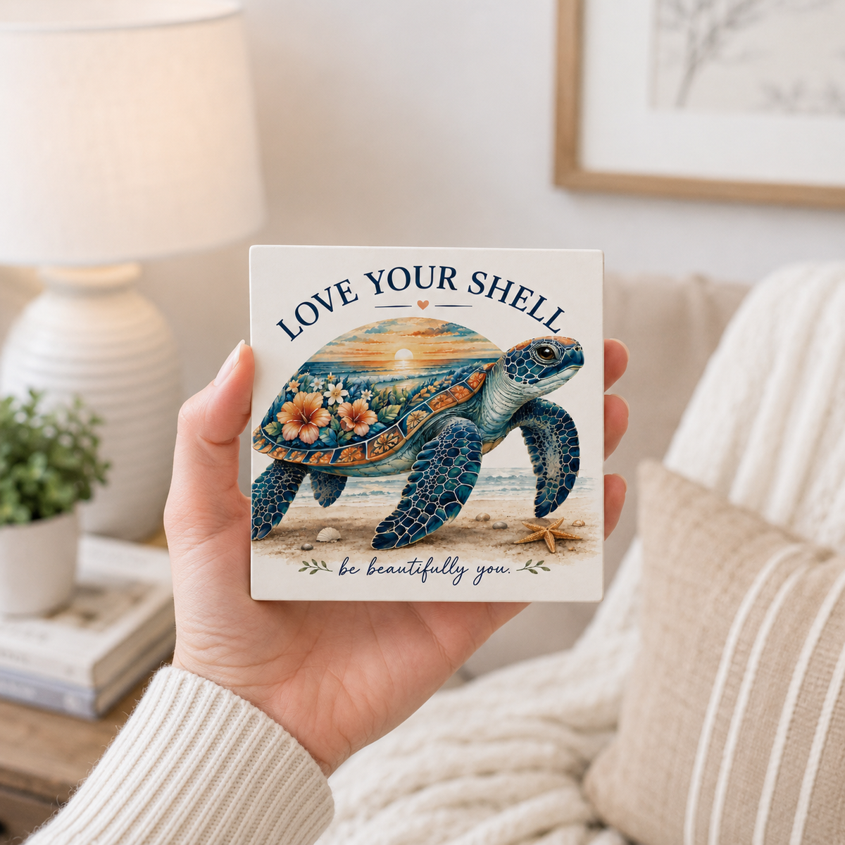 A hand holds a square ceramic tile coaster featuring an illustrated sea turtle with floral patterns and the text 'LOVE YOUR SHELL' and 'be beautifully you'. The background shows a cozy living room setting with a lamp, books, and a striped blanket.