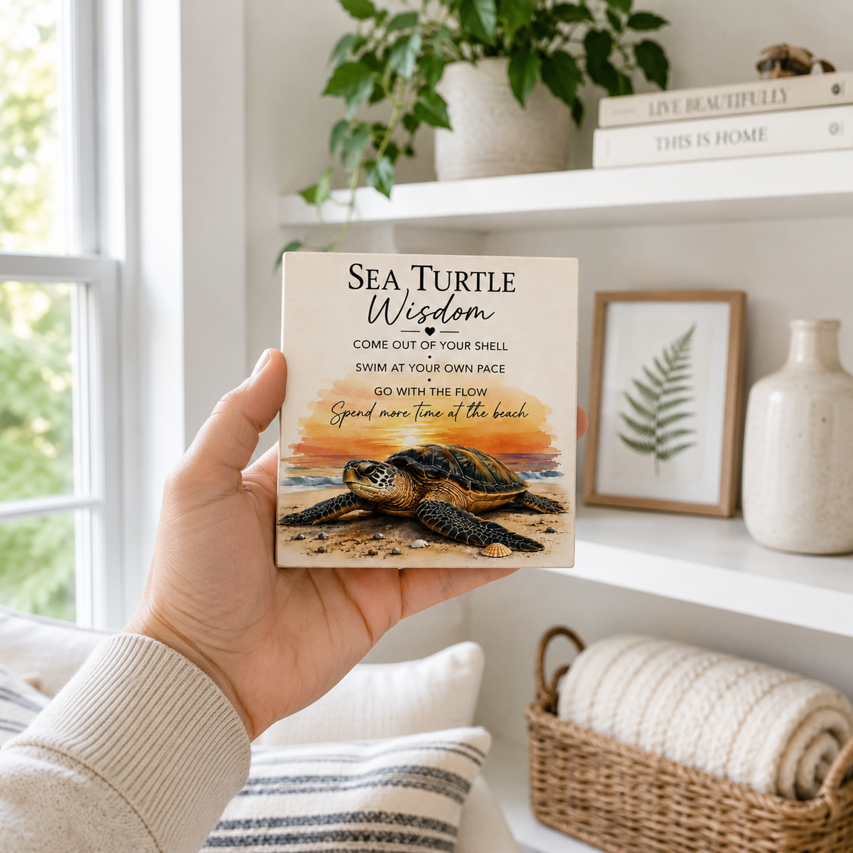 A hand holds a square ceramic tile coaster featuring a sea turtle illustration and the text 'SEA TURTLE Wisdom'. The background shows a cozy, sunlit living room with shelves, a potted plant, and a woven basket.