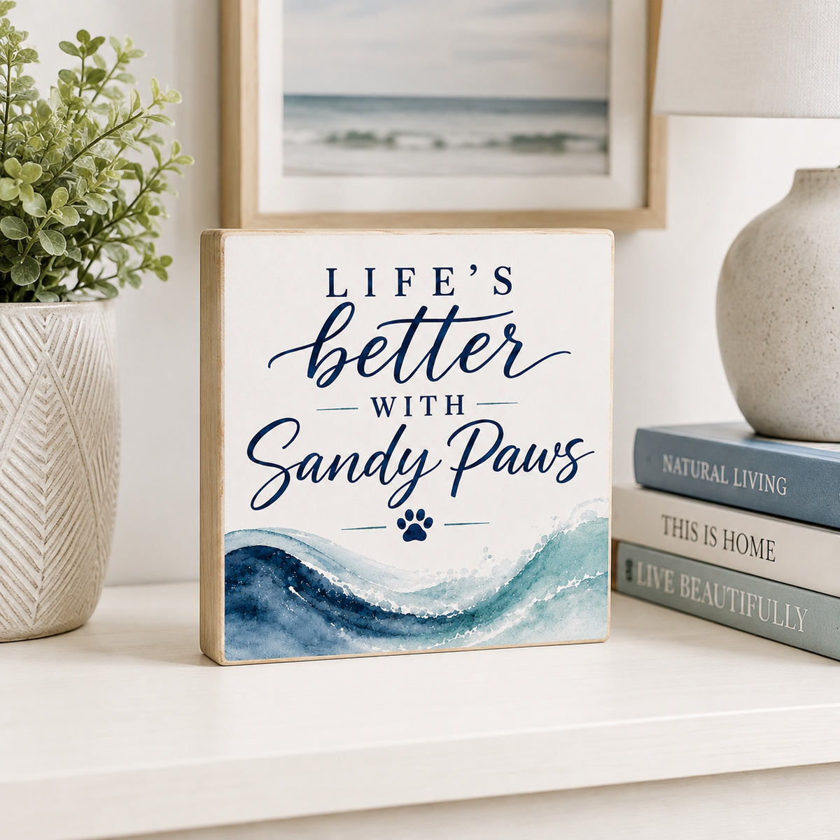 A square decorative sign with the phrase “LIFE’S better WITH Sandy Paws” in navy lettering above a blue watercolor wave design and a small paw print. The sign is styled on a white shelf beside a potted plant, framed art, a lamp, and stacked books.