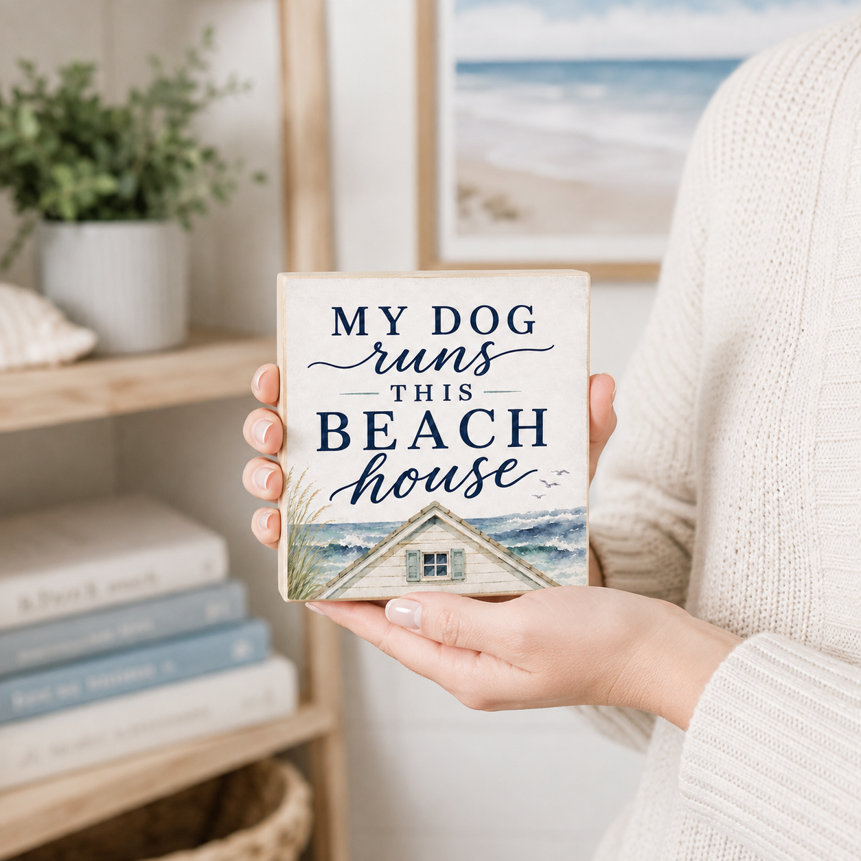 A person in a cream knit sweater holds a square decorative sign that reads “MY DOG runs THIS BEACH house” above a small beach cabin illustration. The background shows a softly blurred beach-themed interior with shelves, books, and a framed ocean scene.
