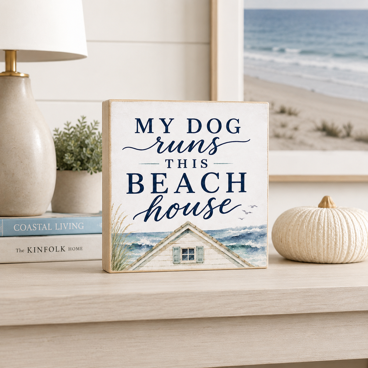 A square decorative sign on a light wood shelf reads “MY DOG runs THIS BEACH house” in navy lettering above a painted beach house and ocean scene. The sign is styled in a home setting beside a lamp, books, a small plant, and a ribbed cream pumpkin, with a beach scene visible in the background.