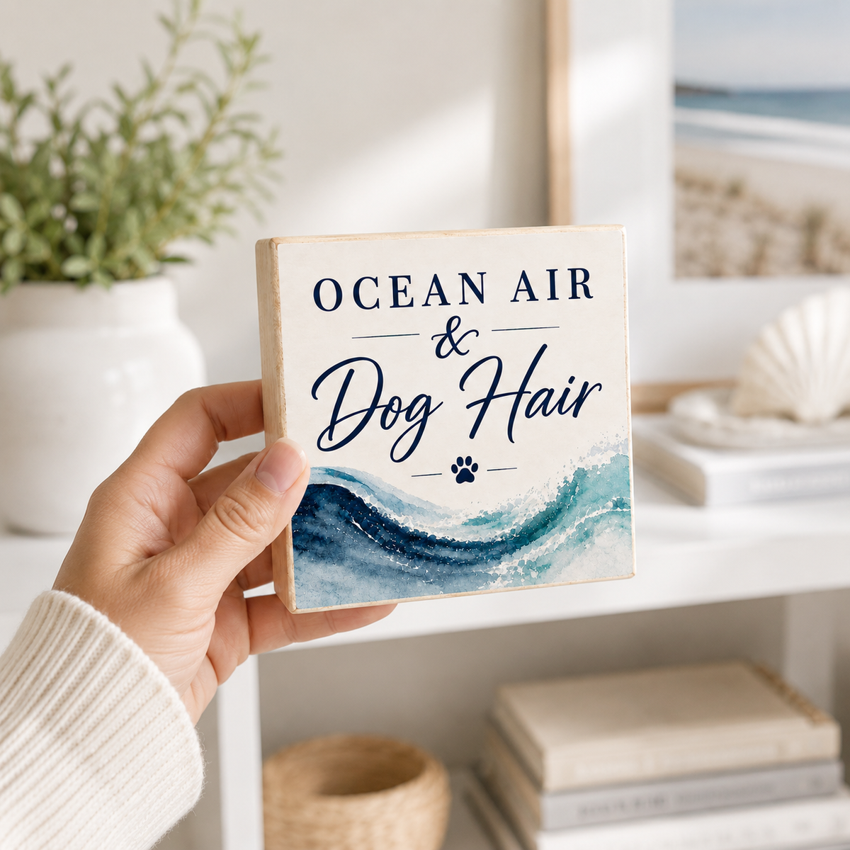 A hand holds a square decorative sign with the text 'OCEAN AIR & Dog Hair' above a blue water wave illustration. The sign is shown in a bright, modern room setting with a shelf, books, and a potted plant in the background.