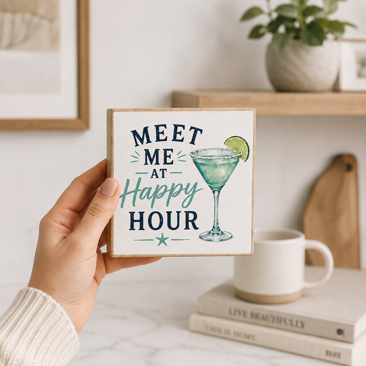 A hand holds a square decorative tile coaster featuring the text 'MEET ME AT Happy HOUR' in navy and teal lettering, alongside an illustration of a martini glass with a lime wedge. The background shows a cozy, styled home interior with a white mug, books, a wooden shelf, and a potted plant.