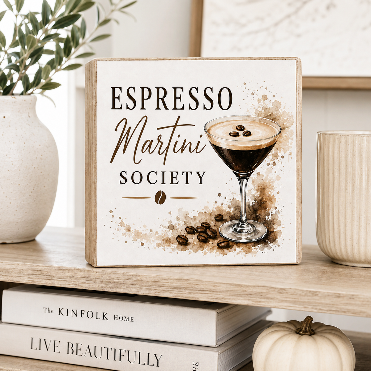 A square decorative sign reading “ESPRESSO Martini SOCIETY” with an illustration of an espresso martini glass and coffee beans, displayed on a wooden shelf with books, a vase, and a small pumpkin.