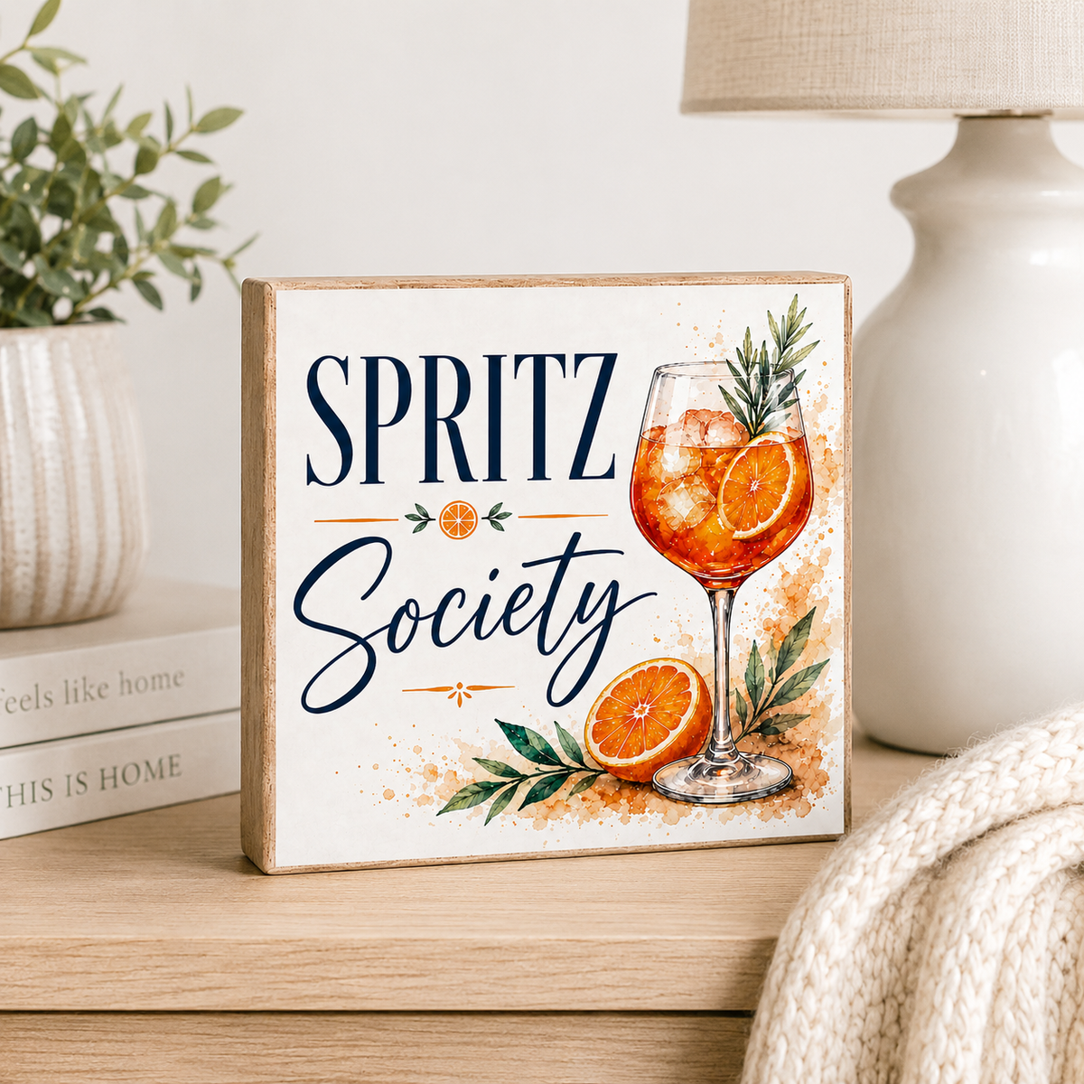 A square decorative wooden sign with a rustic light wood border, featuring the text 'SPRITZ Society' in navy blue script and print, alongside a watercolor illustration of an orange spritz cocktail with a rosemary garnish, orange slices, and green leaves. The sign is styled on a wooden table next to a white ceramic lamp, a small potted plant, and a beige knit blanket.