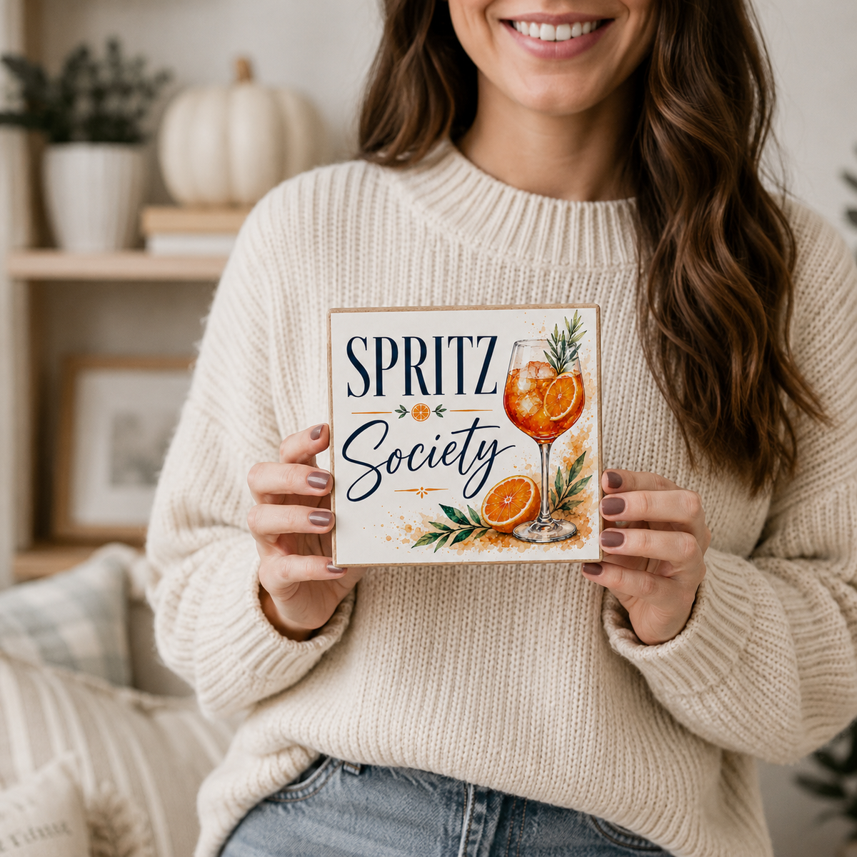 A woman in a cream knit sweater holds a square decorative sign that reads “SPRITZ Society” with an illustrated orange drink and garnishes. The photo is taken in a warm indoor setting with shelves and decor blurred in the background.
